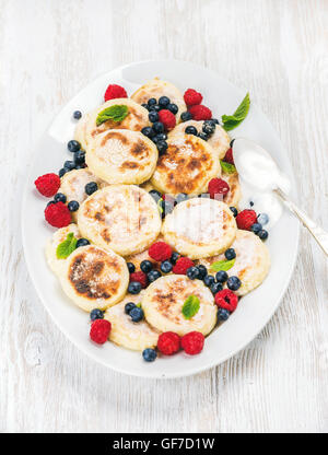 Syrniki o ricotta frittelle con freschi frutti di bosco e panna acida salsa nel piatto di servizio su bianco sullo sfondo di legno Foto Stock