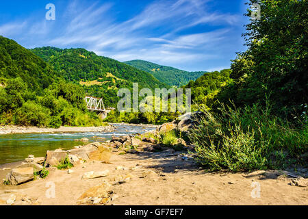 Riva di un fiume di montagna con pietre e ponte di ferro tra la foresta nella zona fural Foto Stock