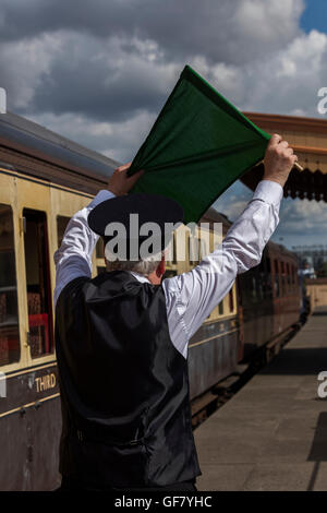 Treno di andana trattiene la sua bandiera verde sulla piattaforma per segnalare al conducente la locomotiva a vapore a lasciare Foto Stock