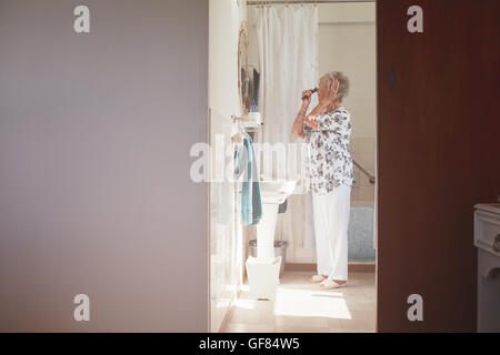 A piena lunghezza shot indoor di senior donna pettinare i capelli a cassettiera. Donna anziana prepararsi in bagno. Foto Stock