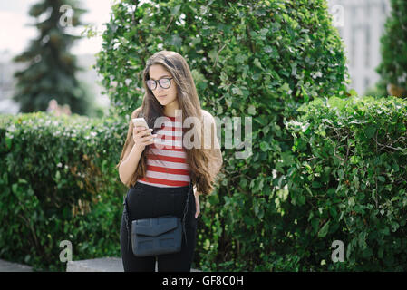 Giovane donna asiatica con gli occhiali utilizza lo smartphone e l'invio di sms. Giovane donna su sfondo della città con foglie verdi. Foto Stock