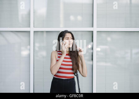 Giovane donna asiatica parlando sullo smartphone vicino a parete in vetro dell'edificio degli uffici. Giovane donna su sfondo della città. Foto Stock