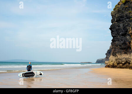 Surfer in piedi con la scheda sulla spiaggia di ballybunion selvatici in modo atlantico Foto Stock