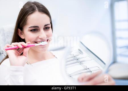 Modello rilasciato. Giovane donna spazzolatura dei denti e guardando la sua riflessione a specchio. Foto Stock