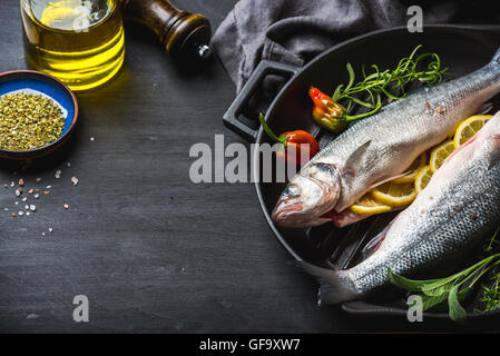 Materie non cotti spigola pesce con erbe e spezie in ghisa pentola di cottura nero su sfondo di legno Foto Stock