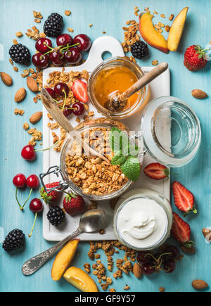 Sana colazione ingredienti. Oat granola in aprire il vasetto di vetro, yogurt, frutta, bacche, miele e menta su sfondo blu Foto Stock