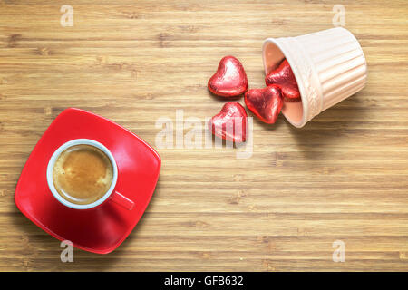 A forma di cuore ad caramelle confezionate in un colore rosso brillante foil giacente sulla texture in legno con tazza di caffè vicino ad essa. Sfondo romantico per i temi. Foto Stock