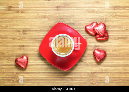 A forma di cuore ad caramelle confezionate in un colore rosso brillante foil giacente sulla texture in legno con tazza di caffè vicino ad essa. Sfondo romantico per i temi. Foto Stock