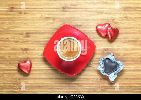 A forma di cuore ad caramelle confezionate in un colore rosso brillante foil giacente sulla texture in legno con tazza di caffè vicino ad essa. Sfondo romantico per i temi. Foto Stock