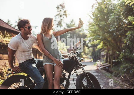 Colpo di coppia giovane tenendo selfie sulla motocicletta. Giovane uomo e donna prendendo ritratto di auto mentre è in moto sulla strada del villaggio. Foto Stock