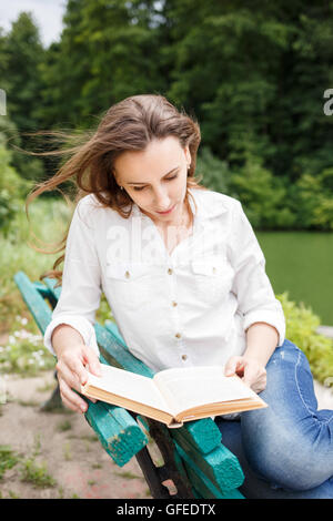 Giovane donna seduta nel parco vicino al laghetto e la lettura del libro. Bella ragazza caucasica con prenota relax sul banco di lavoro Foto Stock