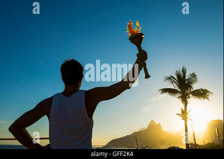 Silhouette di torchbearer atleta in piedi con la torcia di sport di fronte al Rio de Janeiro, Brasile skyline tramonto a Ipanema Foto Stock