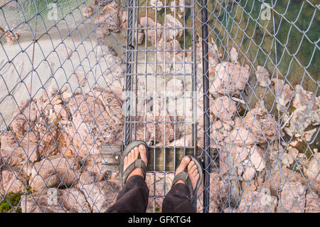 Guardando verso il basso sui piedi in flip-flop su un alto e pericoloso airwalk con un fiume e rocce al di sotto di Foto Stock