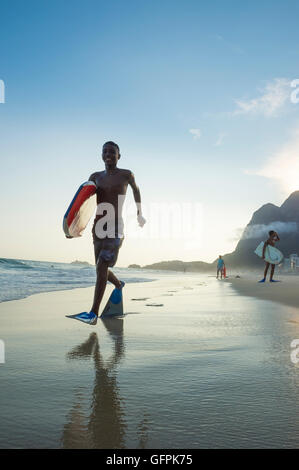 RIO DE JANEIRO - Marzo 8, 2016: Bodyboarders a piedi su São Conrado spiaggia sotto un tramonto della silhouette iconica Pedra da Gávea. Foto Stock