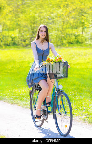 Giovane donna in corto abiti colorati con capelli lunghi cavalca una bicicletta con cestello e fiori tour estate città park, look e sorriso Foto Stock