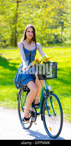 Giovane donna in corto abiti colorati con capelli lunghi cavalca una bicicletta con cestello e fiori tour estate città park, look e sorriso Foto Stock