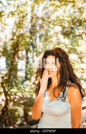 Close up busty elegante donna matura sorridente verso la telecamera mentre copre la bocca con la mano contro il giardino verde con uno sfondo con spazio di copia Foto Stock
