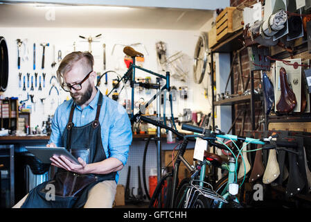 Un uomo che lavora in un negozio di riparazione di biciclette seduto utilizzando una tavoletta digitale Foto Stock