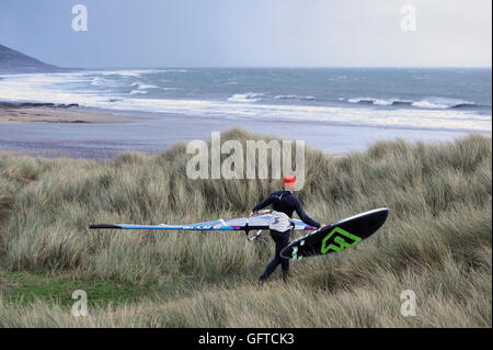 Windsurf controlla le condizioni di surf come egli cammina oltre le dune della spiaggia a Port Eynon, Gower, Galles Foto Stock