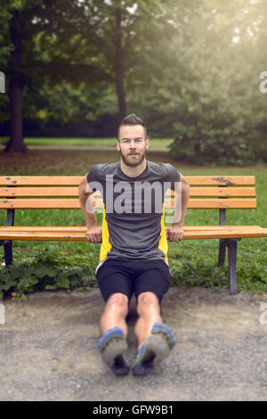 Giovane uomo esercizio utilizzando un di legno una panchina nel parco il sollevamento su se stesso le sue braccia per tonificare i muscoli, in vista frontale con copia spazio in un Foto Stock