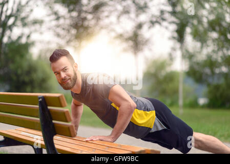 Giovane uomo che lavora fuori facendo push-up su una tavola di legno panchina come egli si riscalda per il suo allenamento quotidiano o jog Foto Stock