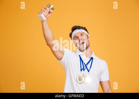 Felice giovane atleta con tre medaglie e trofeo cup celebrando la vittoria su sfondo giallo Foto Stock