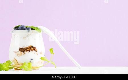 Yogurt oat granola con marmellata di mirtilli e foglie verdi in un barattolo di vetro sul pastello sfondo lilla Foto Stock