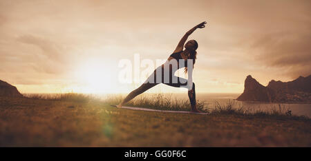 Colpo orizzontale di donna sana che pratica Utthita Parsvakonasana yoga asana. donna in forma fisica che si esercita sulla scogliera durante il sole Foto Stock