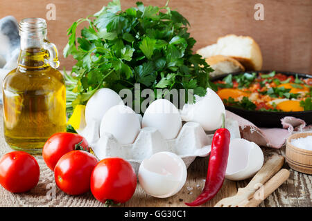 Uova, pomodori, olio d'oliva, prezzemolo, peperoncino. Ingredienti per shakshuka su tavola in legno rustico Foto Stock