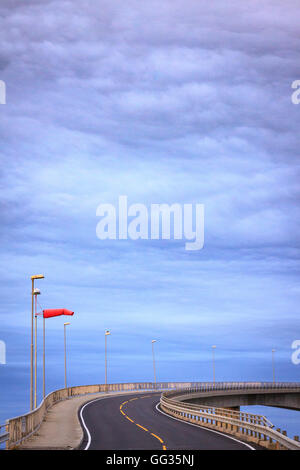 La curvatura della strada su un ponte. Indicatore del vento soffia orizzontalmente, indicando i venti pesanti. Nuvola grande consistenza come copia di spazio. Foto Stock