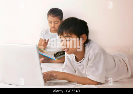 Boy utilizzando computer portatile durante la lettura della ragazza prenota nel letto Foto Stock