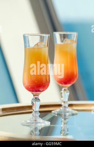 Due bicchieri contenenti Tequila Sunrise orange cocktail drink con ghiaccio su un vetro tavolo accanto a una finestra. Foto Stock