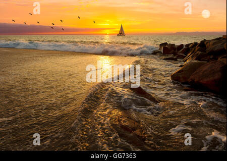 Barca a vela al tramonto è una barca a vela vela in mare contro un vivido arancione tramonto con uno stormo di uccelli in seguito lungo come una luna piena Foto Stock