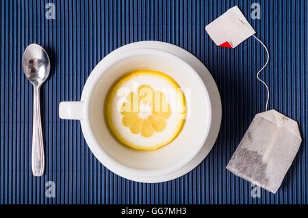 Una composizione semplice con tazza da tè vuota ma con fetta di limone piattino e cucchiaio, sacchetto filtro di tesu blu sullo sfondo a strisce Foto Stock