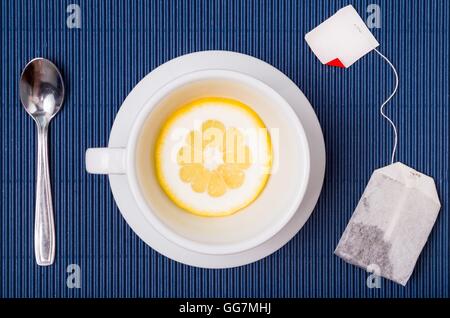 Una composizione semplice con tazza da tè vuota ma con fetta di limone piattino e cucchiaio, sacchetto filtro di tesu blu sullo sfondo a strisce Foto Stock