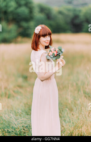 Bella sognante dai capelli rossi sposa con bouquet nuziale si erge sullo sfondo del campo Foto Stock