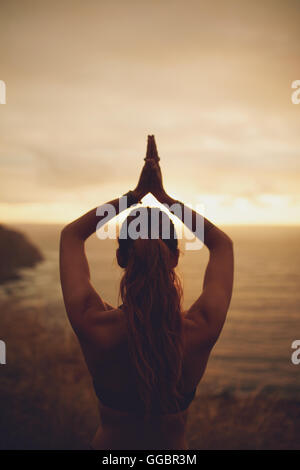 Vista posteriore del colpo di donna fitness fare yoga durante il tramonto. Donna in piedi con le mani giunte overhead. Foto Stock