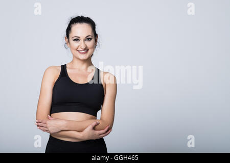 Adatta la giovane donna con le braccia piegate guarda alla fotocamera e sorride alla fotocamera in uno studio fotografico su uno sfondo bianco con spazio di copia a destra. Foto Stock