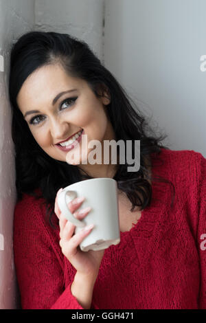 Una donna sorridente e sicura di sé, felice della vita, riposa la testa mentre si siede accanto a una finestra e tiene una bevanda calda in un momento di riflessione Foto Stock