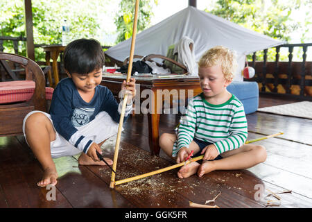 Ragazzo Balinese giocando con il ragazzo straniero, tedesco, 3 anni, bionda, coltello, whittle, carving un bastone di legno, terrazza in legno, Baline Foto Stock