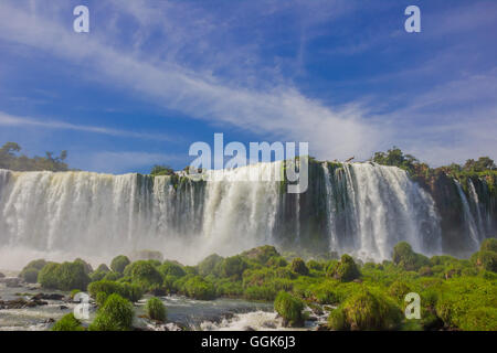 IGUAZU, Brasile - 14 Maggio 2016: bella vista dal fondo delle cascate, alcune rocce ricoperte da erba di fronte alla cascata e il cielo blu come sfondo Foto Stock