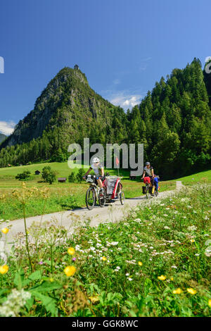 Due ciclisti con rimorchio per bambini equitazione lungo Inn percorso ciclabile, rovina Kronburg in background, Zams, Tirolo, Austria Foto Stock