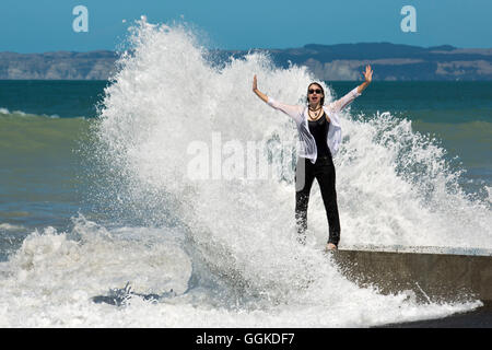 Felice donna in piedi sul mare e ottenere spruzzato da un'onda (MR), Napier, Hawke's Bay, Isola del nord, Nuova Zelanda Foto Stock