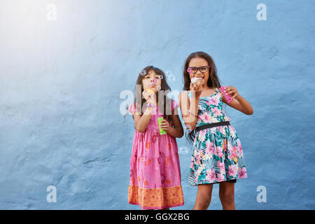 Ritratto di carino bambine soffiando bolle di sapone. Due giovani ragazze giocando contro sfondo blu. Foto Stock