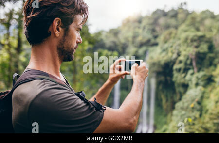 Uomo con zaino in piedi di fronte a cascata e di scattare una foto con il suo telefono cellulare. Escursionista maschio fotografando una caduta di acqua in Foto Stock