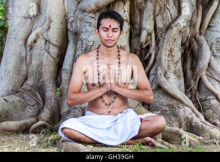 Un giovane uomo indù si siede in meditazione sotto un banyan tree. Foto Stock