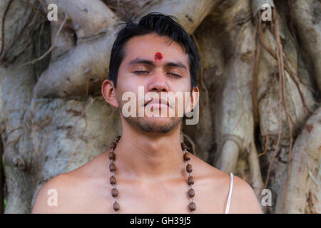 Un giovane uomo indù si siede in meditazione sotto un banyan tree. Foto Stock