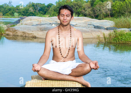 Un giovane sacerdote Indù (bramino), siede in meditazione su una riva di un fiume. Foto Stock