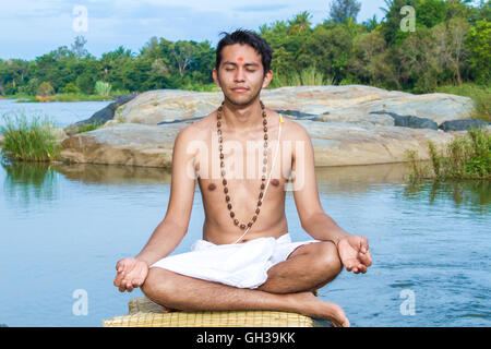 Un giovane sacerdote Indù (bramino), siede in meditazione su una riva di un fiume. Foto Stock