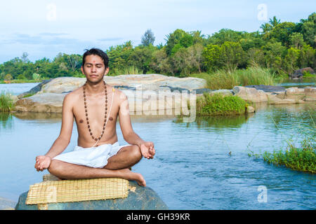 Un giovane sacerdote Indù (bramino), siede in meditazione su una riva di un fiume. Foto Stock